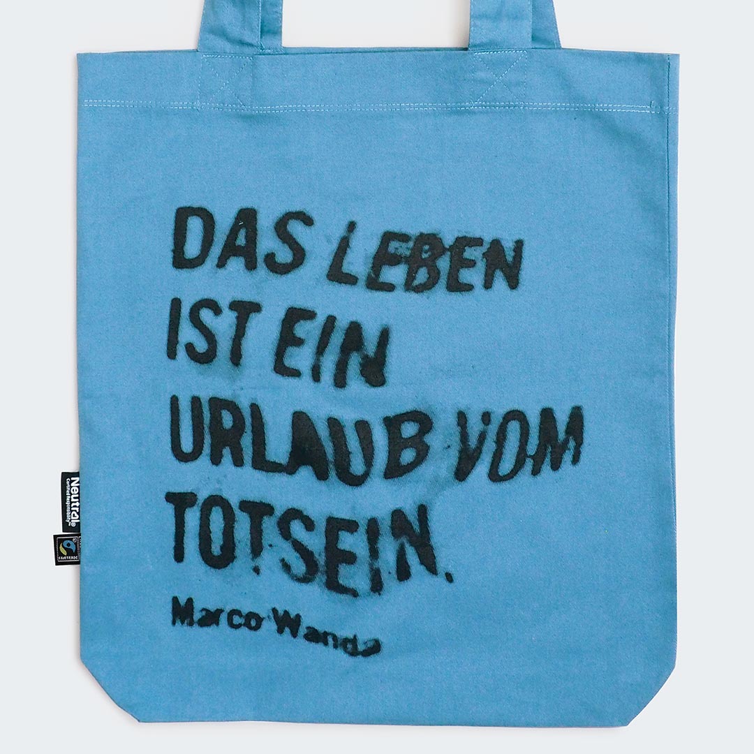 Auf dem Bild ist eine hellblaue Stofftasche mit zwei Henkeln zu sehen. In schwarzer, leicht verwischter und unregelmäßiger Schrift steht auf der Vorderseite der Tasche der Spruch: „DAS LEBEN IST EIN URLAUB VOM TOTSEIN.“ Darunter steht der Name Marco Wanda als Zitatgeber.