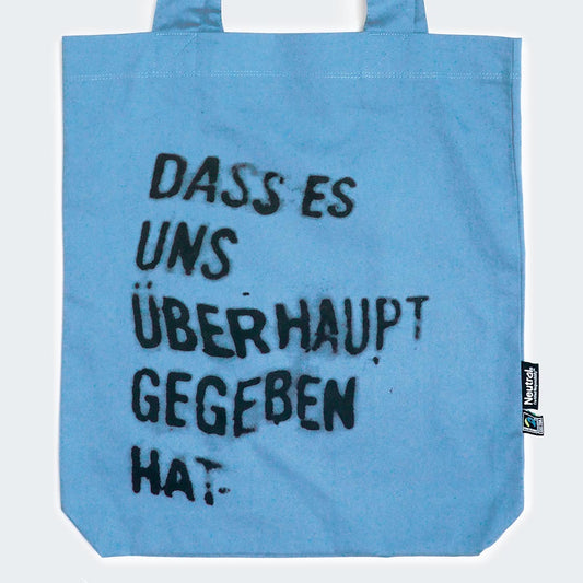 Auf dem Bild ist eine hellblaue Stofftasche mit zwei Henkeln zu sehen. In schwarzer, leicht verwischter und unregelmäßiger Schrift steht auf der Vorderseite der Tasche der Spruch: „DASS ES UNS ÜBERHAUPT GEGEBEN HAT."