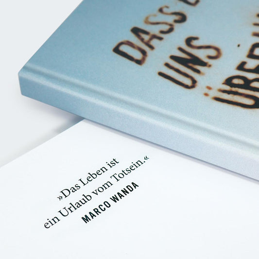 Auf dem Bild ist ein Ausschnitt der Vorderseite des Notizbuches sowie einer Buchinnenseite zu sehen. Der obere Teil zeigt eine Nahaufnahme des Buchdeckels, der einen sanften Farbverlauf von Hellblau zu Beige aufweist. Darauf ist in großer, rostbrauner, leicht verwitterter Schrift der Text zu lesen:
„DASS ES UNS“…
Darunter liegt die erste Seite des Buches, auf der ein Zitat in klarer schwarzer Schrift steht:
„Das Leben ist ein Urlaub vom Totsein.“
Darunter in Großbuchstaben der Name des Zitatgebers: MARCO WA