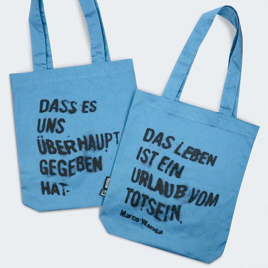 Auf dem Bild sind die Vorder- und Rückseite einer hellblauen Stofftasche zu sehen. Die Tasche hat zwei lange Tragehenkel und weist auf Vorder- und Rückseite unterschiedliche Sprüche von Marco Wanda auf:
Die Vorderseite trägt den Text:
„DASS ES UNS ÜBERHAUPT GEGEBEN HAT.“
Die Rückseite trägt den Text:
„DAS LEBEN IST EIN URLAUB VOM TOTSEIN.“
Darunter steht der Name Marco Wanda.