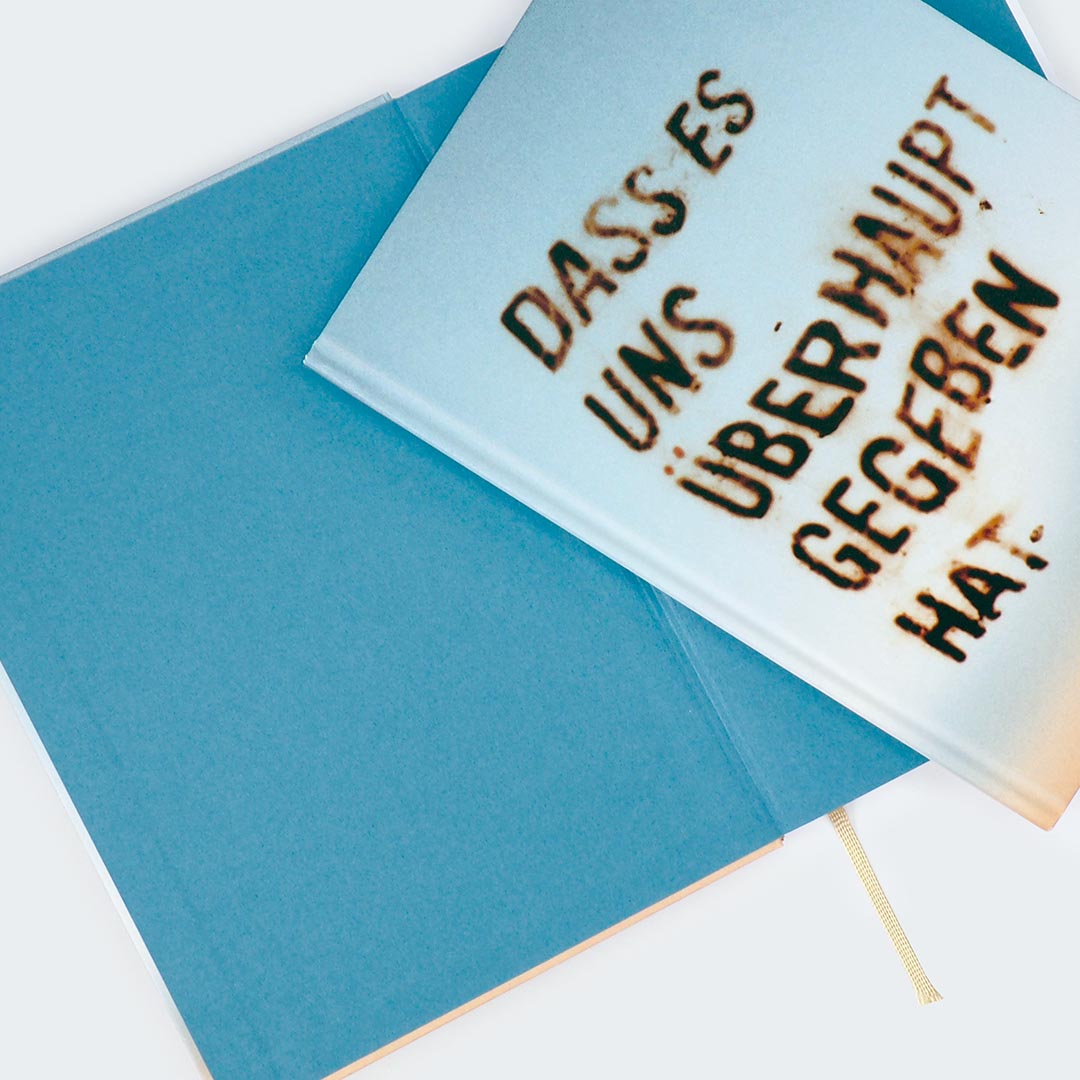 Das Foto zeigt zwei Notizbücher, die teilweise übereinander liegen. Das obere Buch ist leicht schräg positioniert und zeigt das Cover mit einem Farbverlauf von Beige zu Hellblau. Auf dem Cover steht in einer rostbraunen, Schrift: „DASS ES UNS ÜBERHAUPT GEGEBEN HAT.“
Darunter liegt ein geöffnetes Notizbuch mit einer einfarbig blauen Innenseite, der Vorsatz des Buches. Am unteren Rand ist ein hellgelbes Lesebändchen sichtbar, das aus dem Buch herausragt.